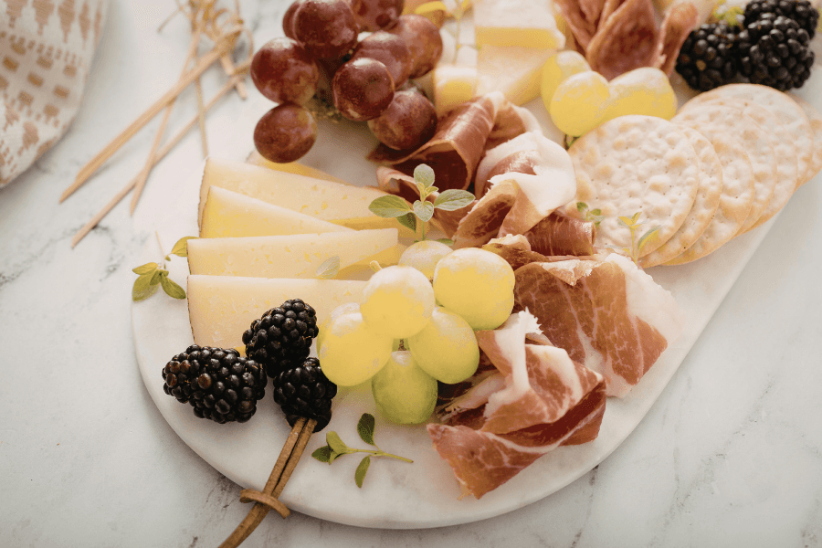 A marble platter filled with assorted cheeses, grapes, blackberries, prosciutto, and crackers.