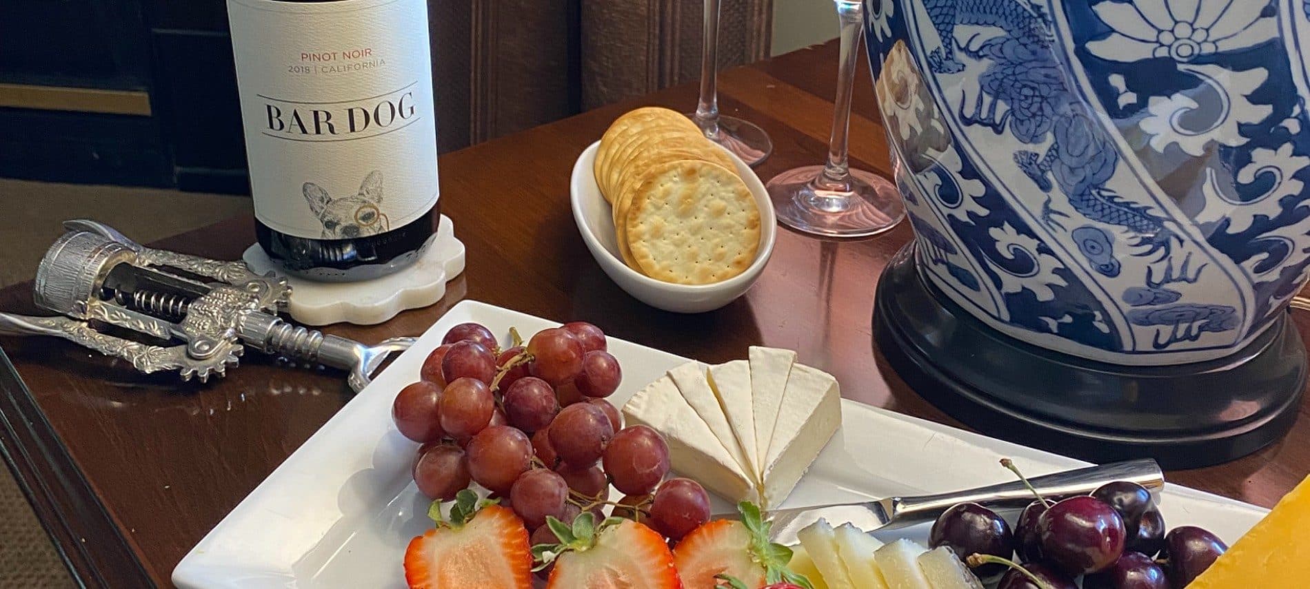 A plate of assorted fruits and cheeses next to a bottle of wine and crackers on a wooden table.
