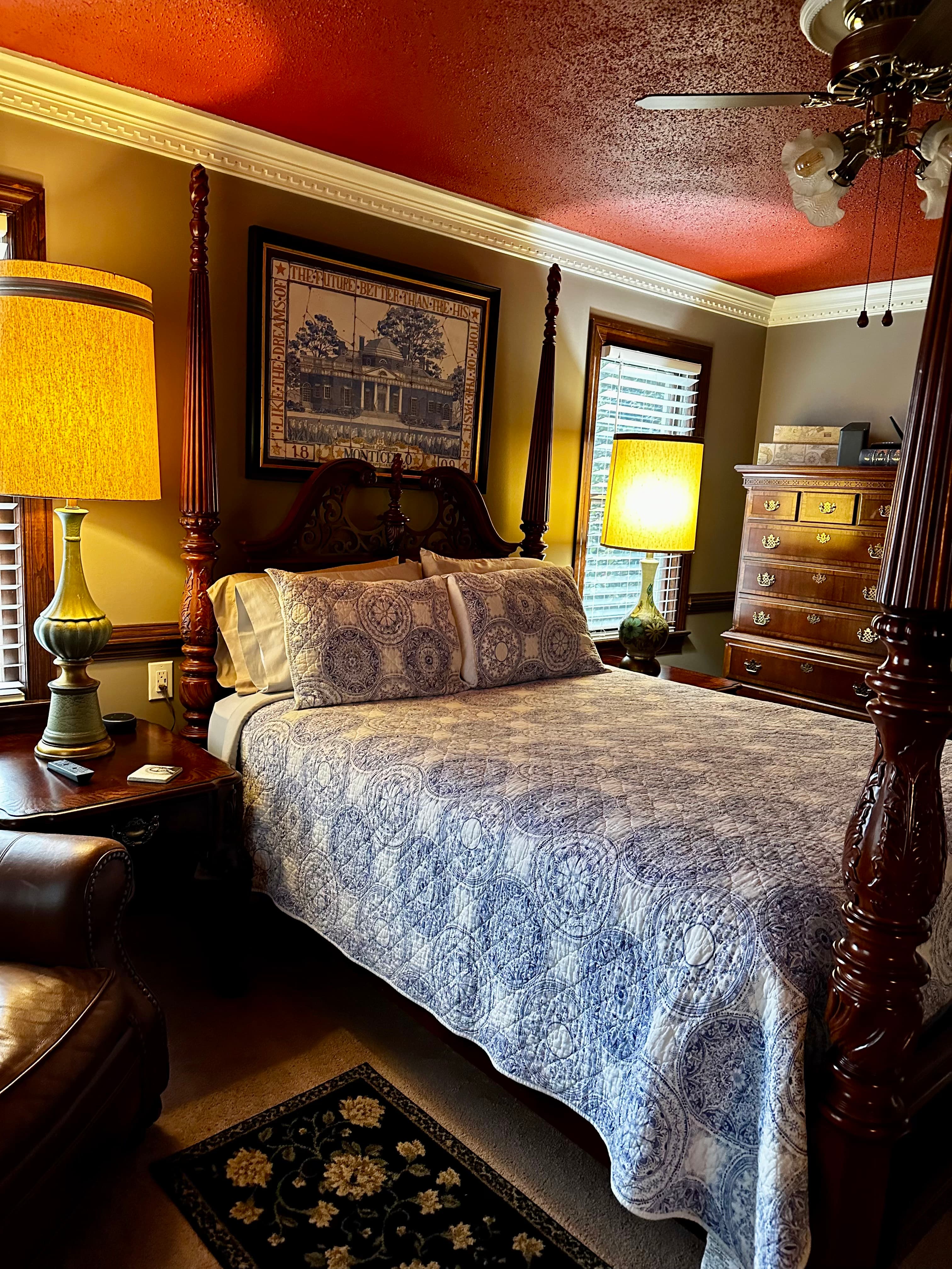 A cozy bedroom featuring a four-poster bed with a blue patterned quilt, warm lighting, and wooden furniture.
