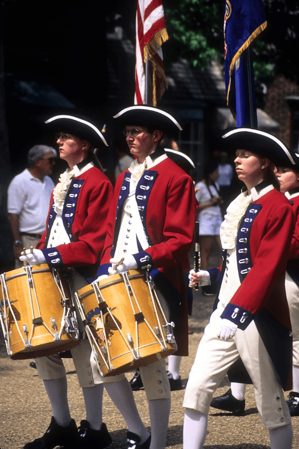 Three musicians in 18th-century Revolutionary War uniforms march in a parade, carrying drums and a flag.