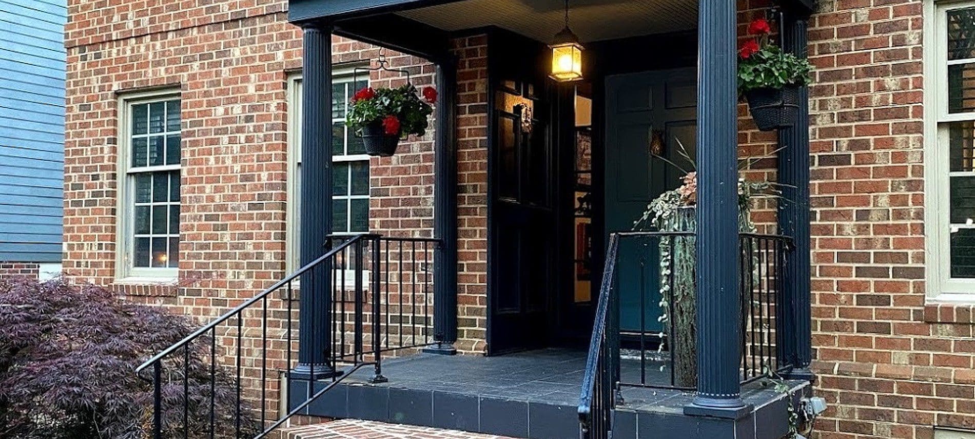 Front porch of a brick home with tall windows and hanging flower plants