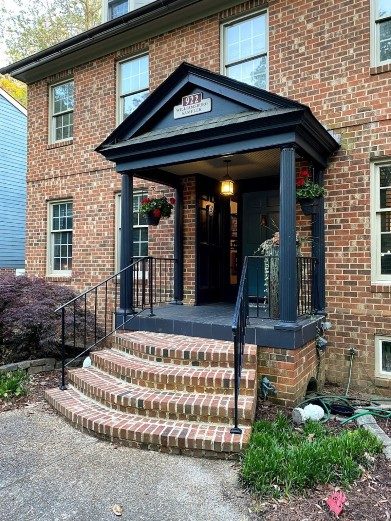 Front porch of a brick home with tall windows, hanging flower plants and steps down to a sidewalk