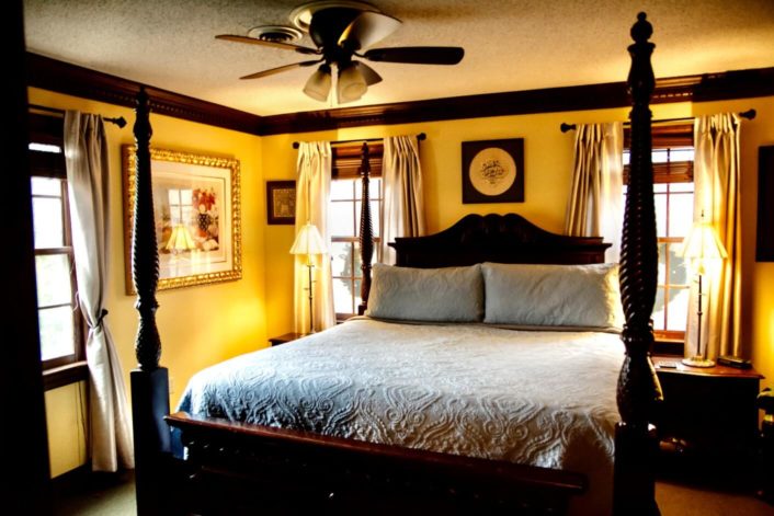 Bedroom with dark wood accents, four poster bed and three bright windows with curtains