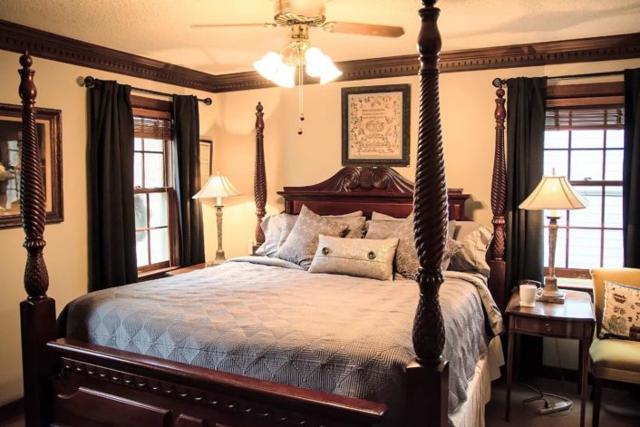 Bedroom with dark wood accents, four poster bed and two bright windows with curtains